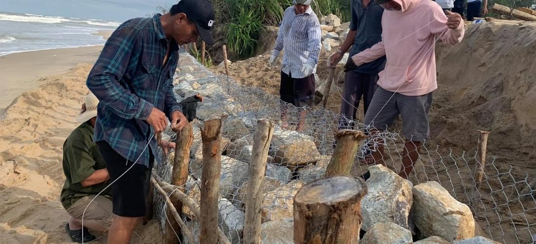 Mô tả chi tiết cảnh xếp chồng rọ đá trong một công trình chắn sóng lớn tại Việt Nam, với các khối rọ đá được xếp lớp cẩn thận dọc theo bờ biển dưới bầu trời nhiều mây.